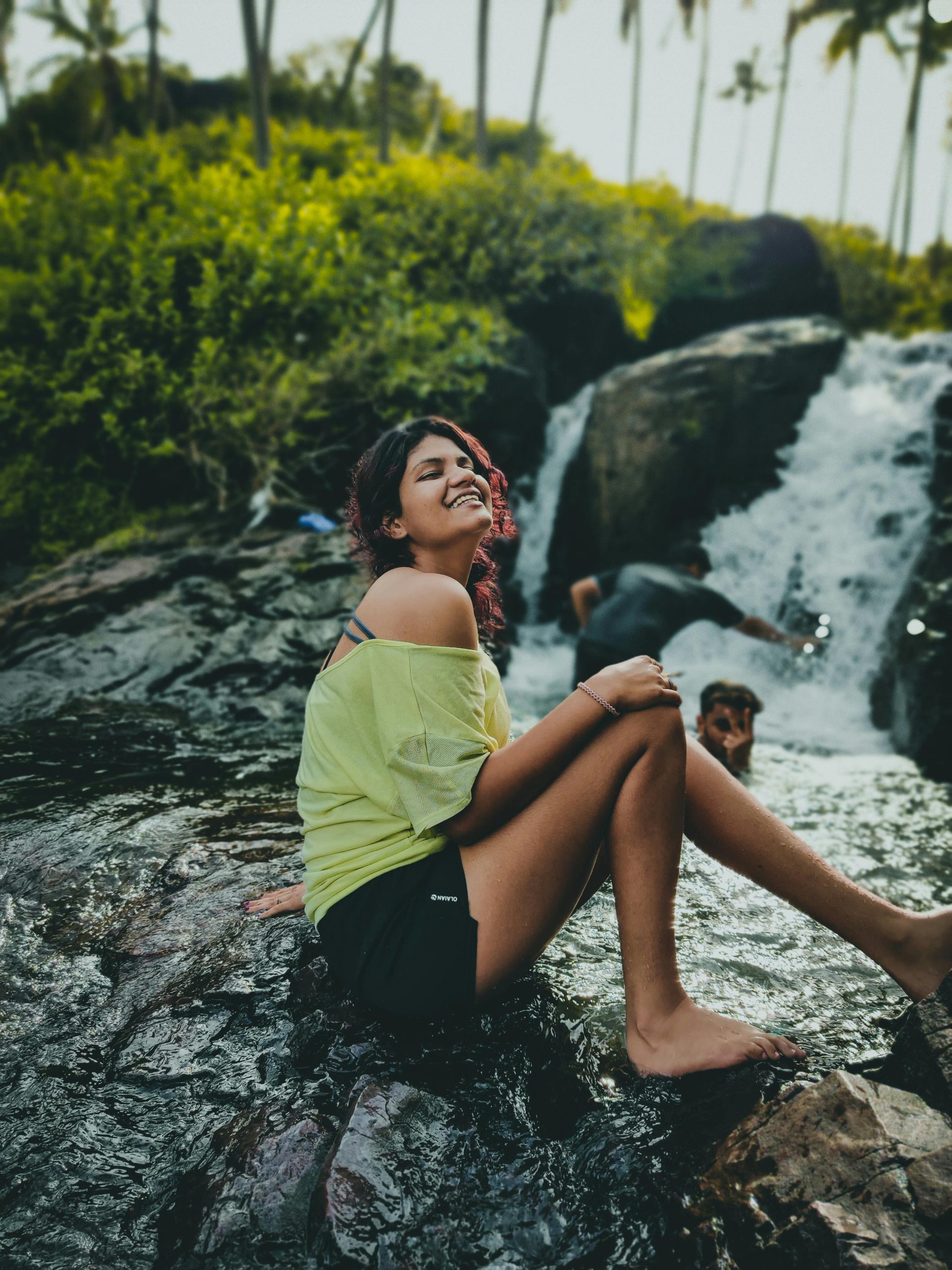 A woman enjoys the summer by sitting on rocks near a cascading waterfall, showcasing happiness and relaxation.