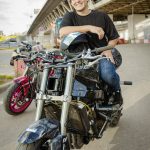 Smiling man sitting on a motorcycle under an urban bridge, enjoying a relaxed moment.